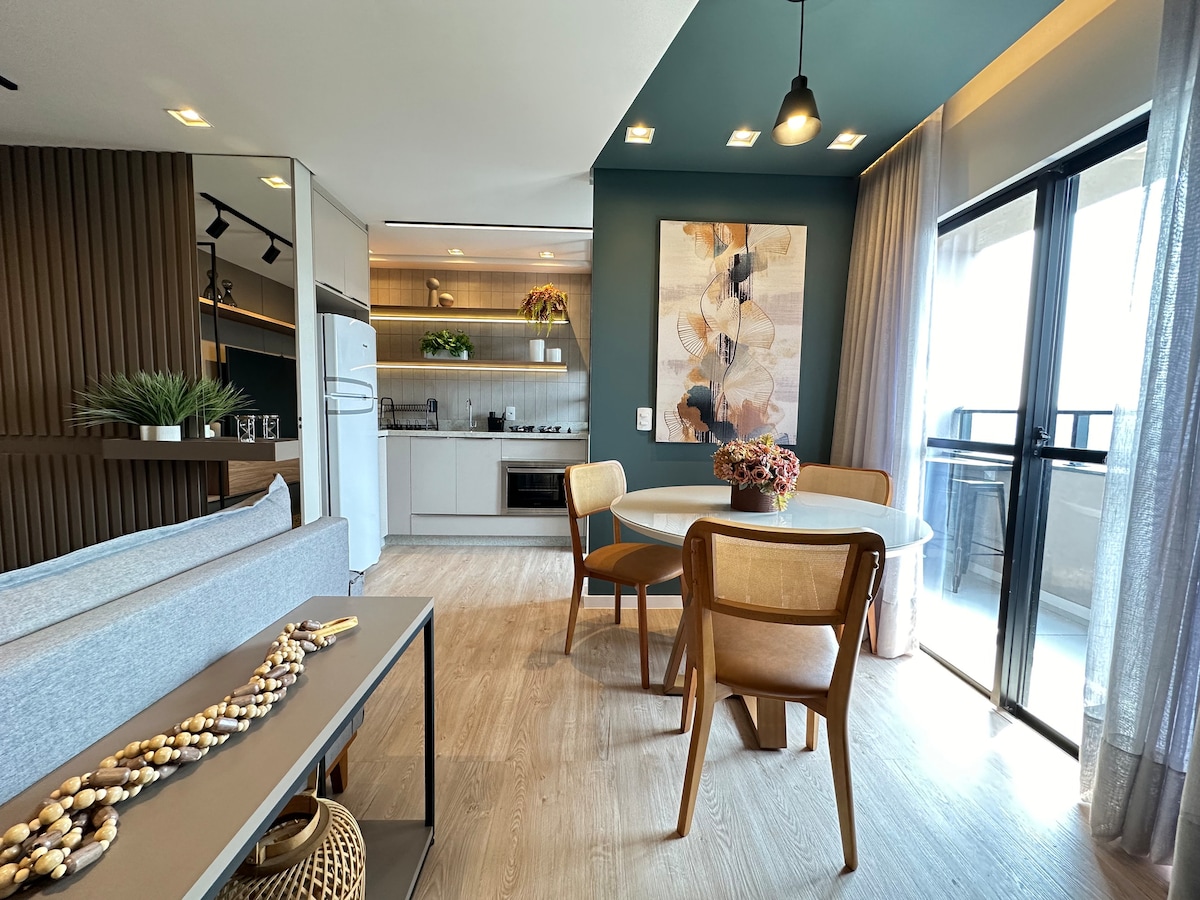 The open-plan living area features a modern kitchen with sleek cabinetry and appliances. A round dining table with wooden chairs is situated near large glass doors leading to a balcony. Natural light enhances the space, complemented by decorative elements like a vase of flowers.