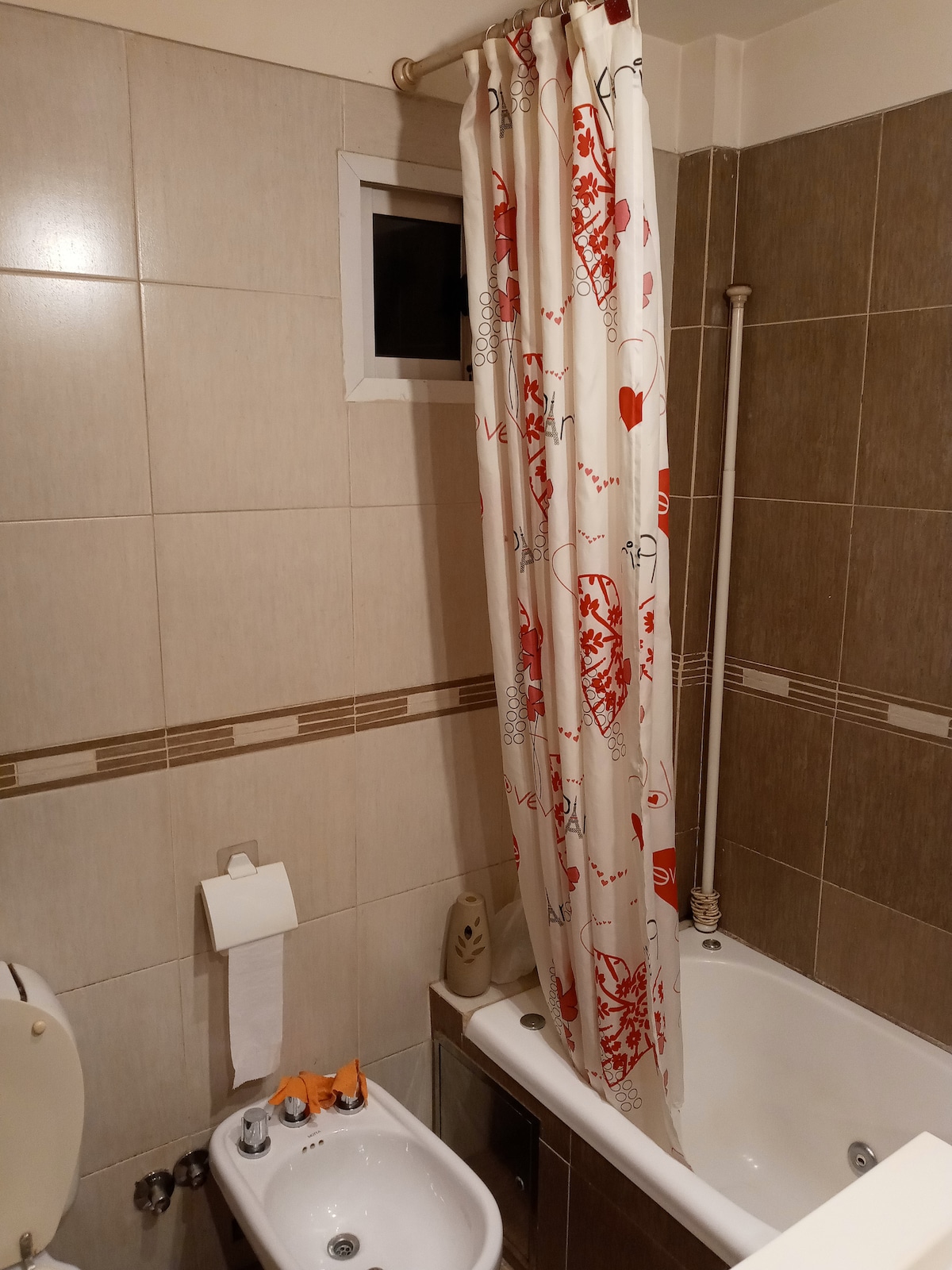 The bathroom features neutral-toned tiles with a bathtub equipped with a showerhead. A decorative curtain with a simple design hangs by the bathtub. A bidet and a wall-mounted toilet are present, along with a toilet paper holder and a small decorative item.