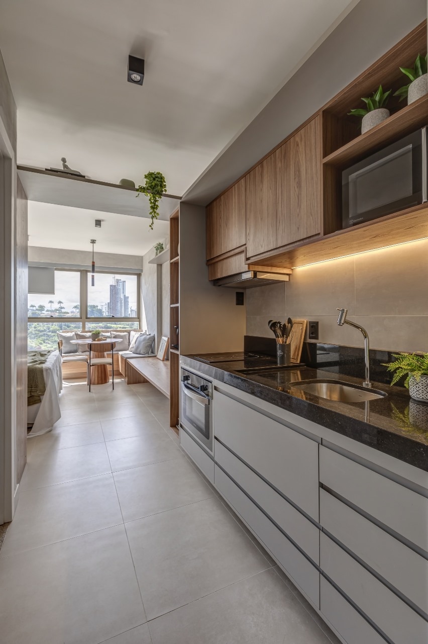 A modern kitchen features sleek cabinets and a dark countertop, complemented by plants for a touch of greenery. Natural light floods through a nearby window, illuminating the cooking area, which includes a stove, sink, and essential kitchen appliances.
