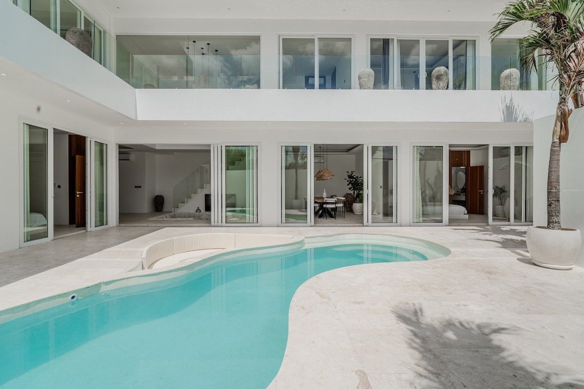 An elegant villa showcases a curved swimming pool surrounded by a spacious outdoor area. Large glass doors and windows reflect natural light, enhancing the open layout. Lush greenery and stylish decor elements add a touch of warmth to the serene ambiance.