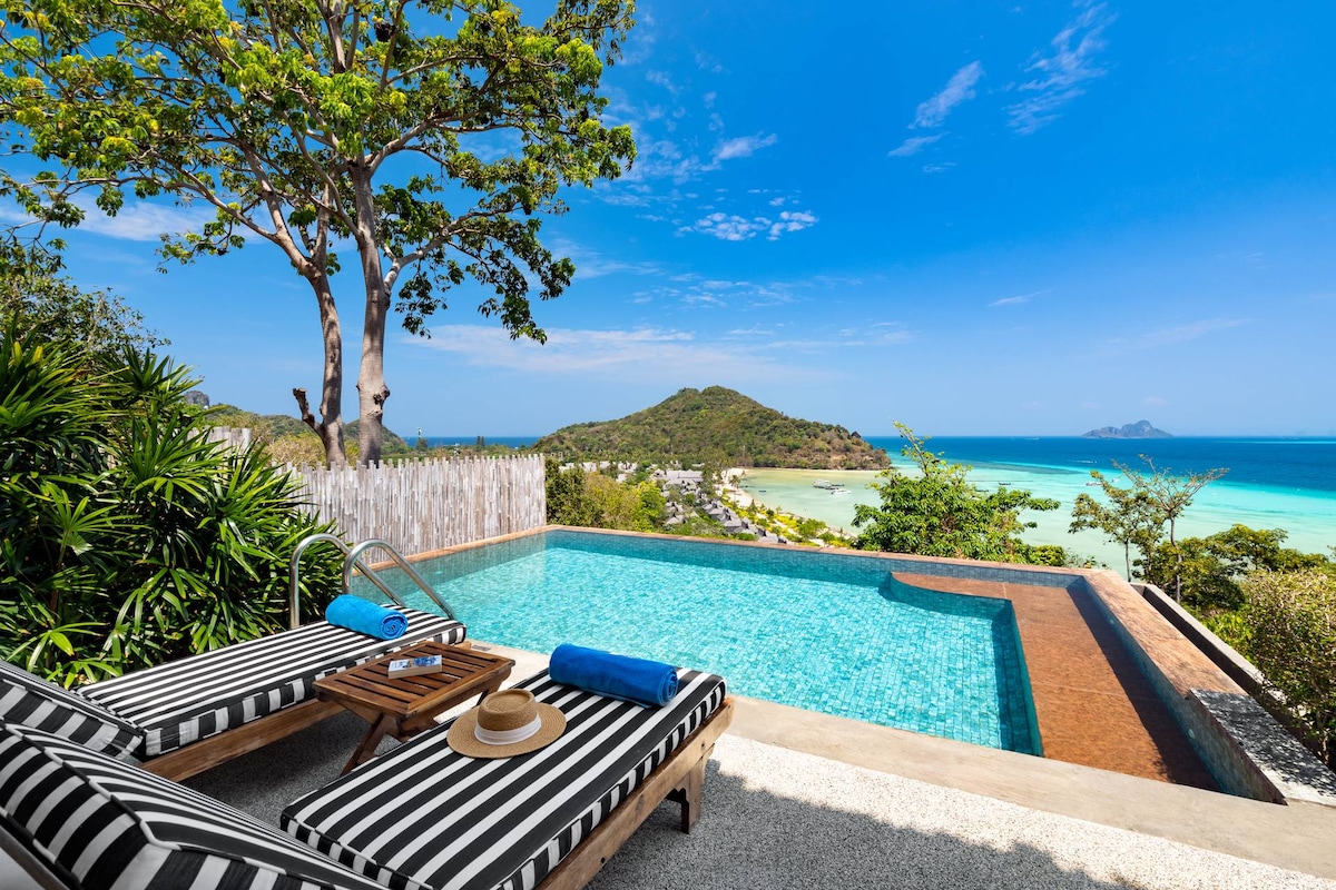 A private infinity pool overlooks the ocean and distant islands, surrounded by lush greenery. Two striped lounge chairs are positioned beside the pool, accompanied by a small side table. Bright blue towels rest on the chairs, enhancing the inviting outdoor space.