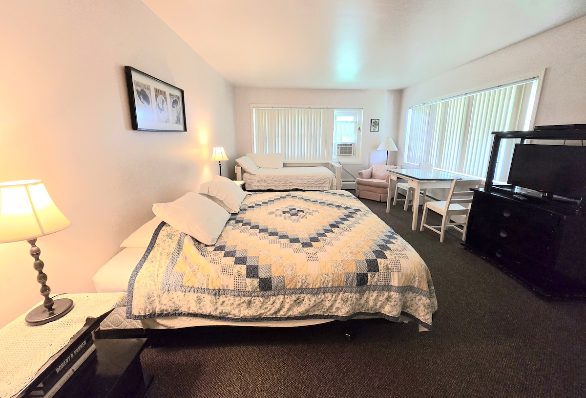 A cozy room features two beds with patterned quilts, enhancing the comfortable sleeping space. A small dining area is visible with a table and chairs, while large windows allow natural light. A television and air conditioning unit add convenience to the setting.