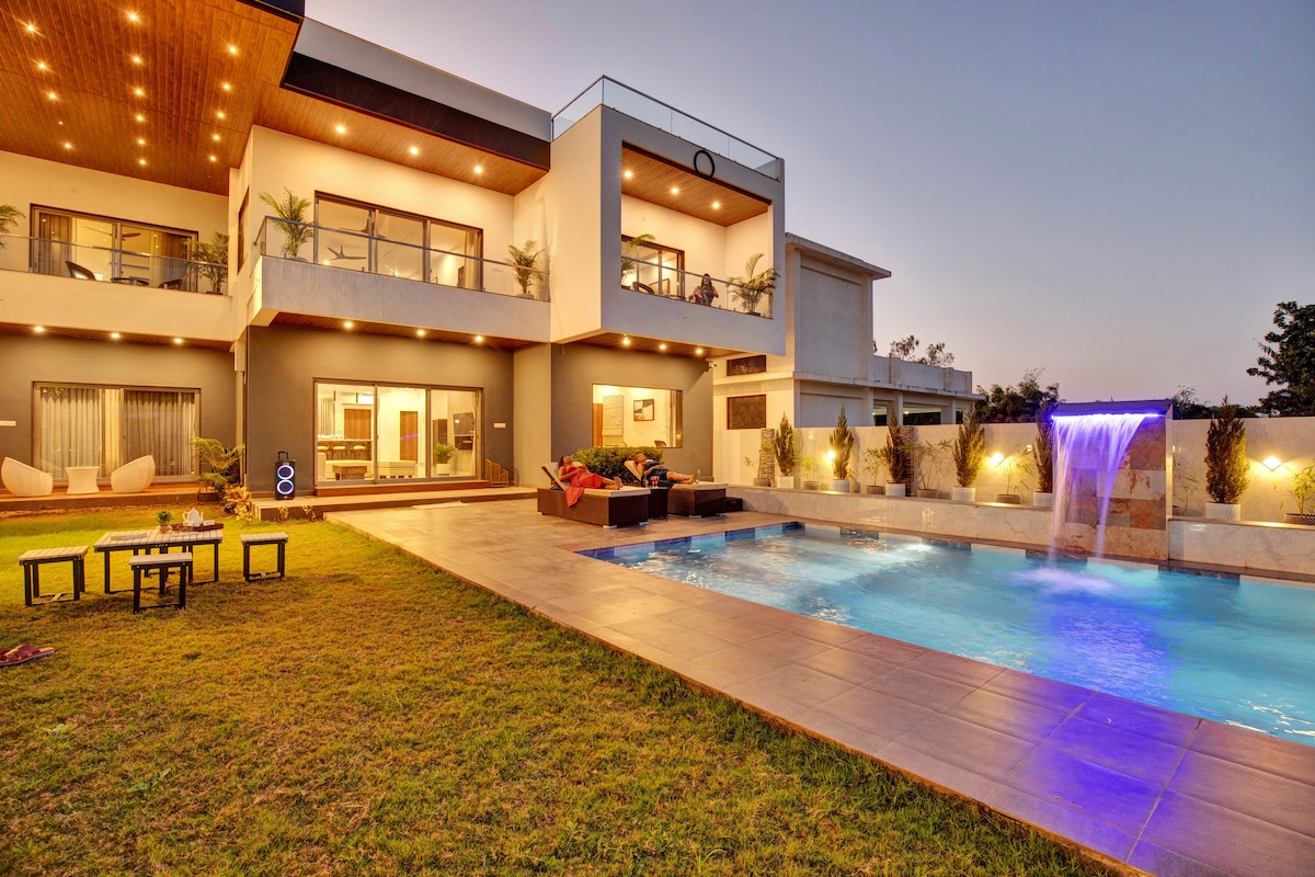 The villa features an outdoor swimming pool illuminated with soft blue lights, accompanied by a playful water fountain. A spacious lawn includes seating and a table, while the modern architecture showcases large windows and a balcony adorned with greenery.