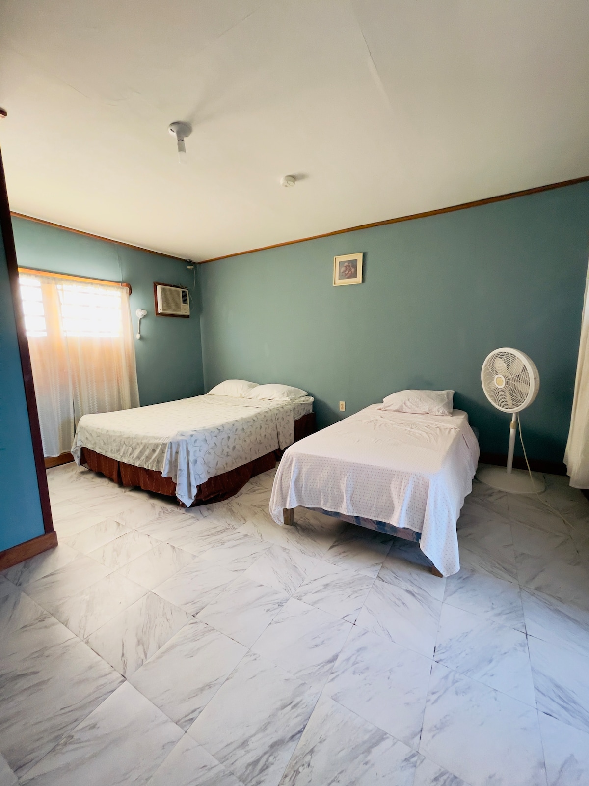 A calming sleeping area features two beds, one with a patterned cover and the other with a plain white sheet. A portable fan is positioned nearby, complementing the natural light that filters through the sheer curtains covering the window.