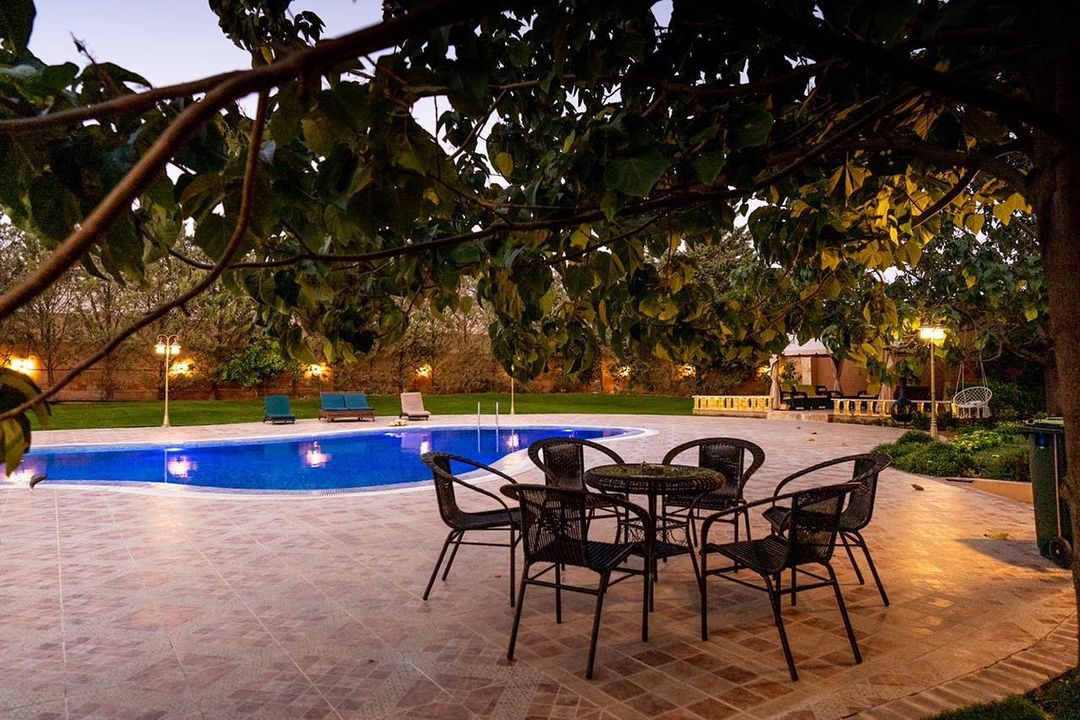 A serene outdoor space features a blue private pool surrounded by lounge chairs and a shaded seating area with a circular table and six woven chairs. Soft lighting from surrounding fixtures enhances the peaceful ambiance, while the landscaped garden adds to the natural beauty.