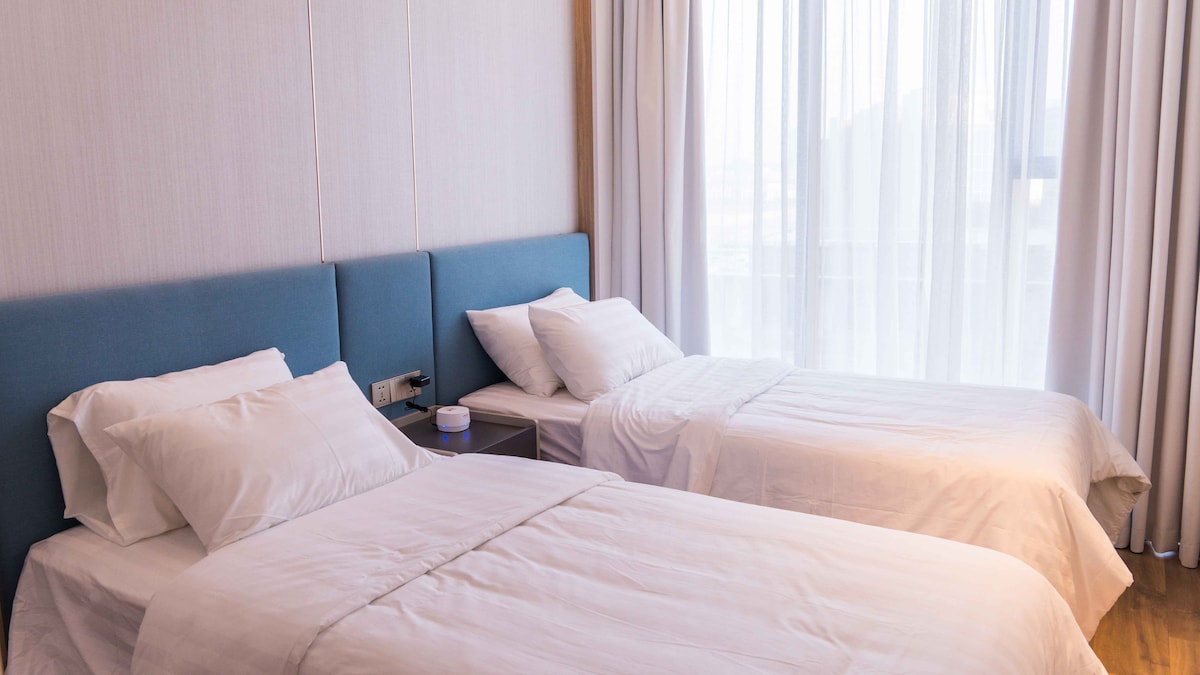 The sleeping area features two spacious beds with light bedding, positioned parallel to large windows that allow natural light to fill the room. Soft curtains frame the windows, and a bedside table is visible next to one of the beds.