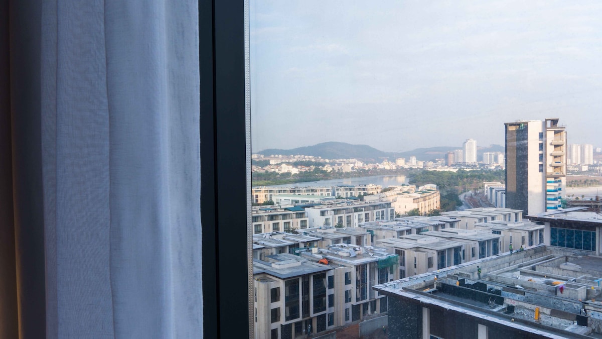 A view from the window frames a cityscape featuring modern buildings and lush hills in the background. Soft natural light filters through sheer curtains, enhancing the serene atmosphere of the surroundings.