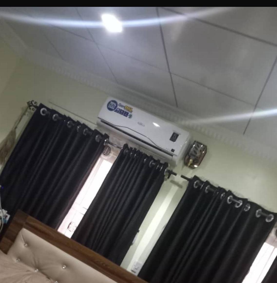 A bedroom features a spacious bed with a wooden headboard, framed by dark curtains. Above, a split air conditioning unit is mounted on the wall, providing cooling comfort. Soft lighting highlights the room's neutral-colored walls and ceiling.