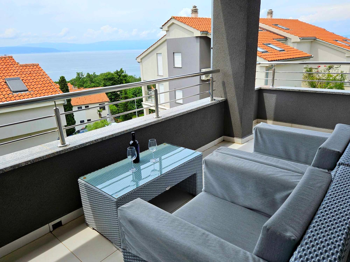 A contemporary outdoor seating area features two gray lounge chairs and a glass-top table. The space is positioned on a balcony, offering views of the sea and surrounding rooftops. The railing is metallic, enhancing the open feel of the terrace.