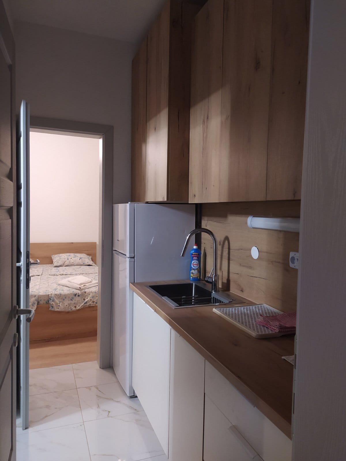 A compact kitchen is presented with wooden cabinetry and a modern sink. A refrigerator stands alongside, with light-colored countertops enhancing the space. A doorway at the far end leads to a bedroom, suggesting a separation between cooking and sleeping areas.