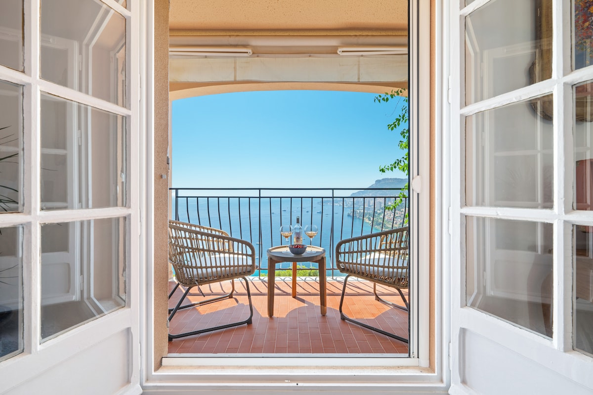 A view from the apartment’s terrace is presented, featuring a small table and two chairs. The railing frames the stunning sight of the Mediterranean Sea and the distant coastline, emphasizing the serene outdoor space accessed through large glass doors.