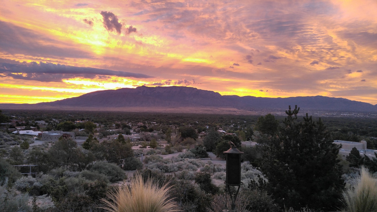 A panoramic view captures the landscape at sunset, with colorful skies illuminated in shades of orange and purple. The distant mountains provide a stunning backdrop against the sprawling valley and scattered homes, surrounded by lush vegetation and trees.
