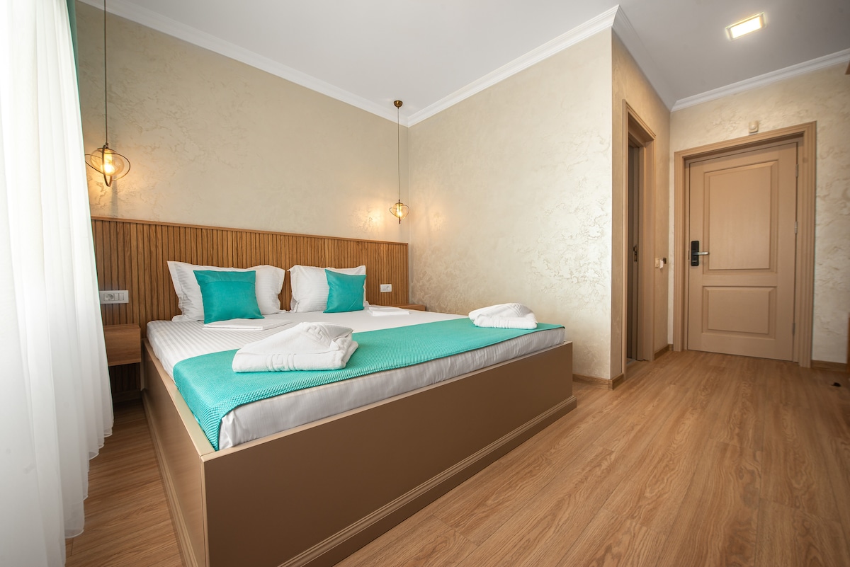 A bright and airy bedroom features a spacious bed adorned with white linens and teal accent pillows. Soft, natural light filters through sheer curtains, illuminating the warm wooden floor. The decor is simple yet elegant, complemented by tasteful lighting fixtures and a wooden accent wall.