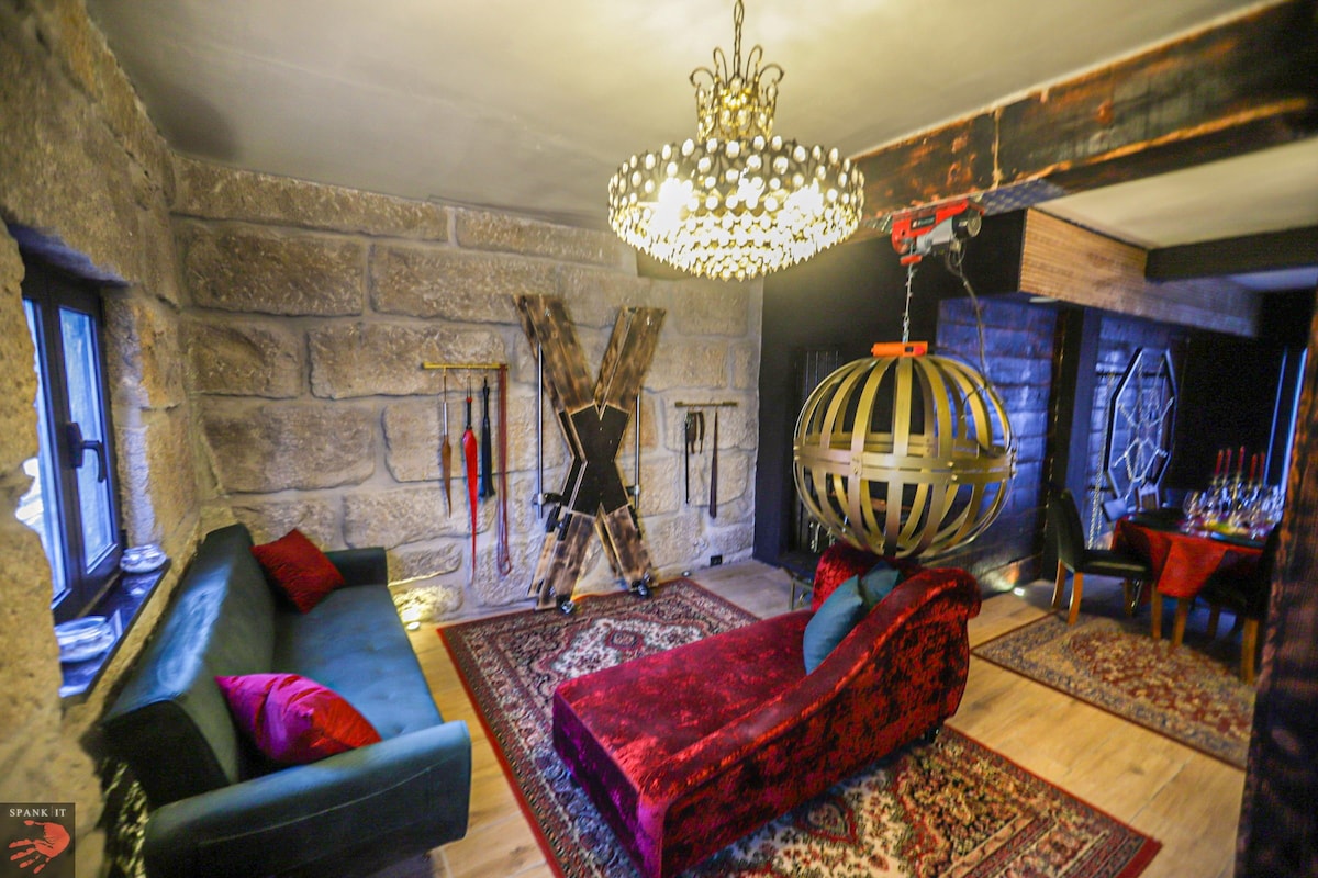 An elegantly furnished room features a striking chandelier and a large round hanging fixture. A red chaise lounge is positioned alongside a dark green sofa, both on a patterned area rug. Various decorative elements and a dining area with a table set for multiple serve as the backdrop.