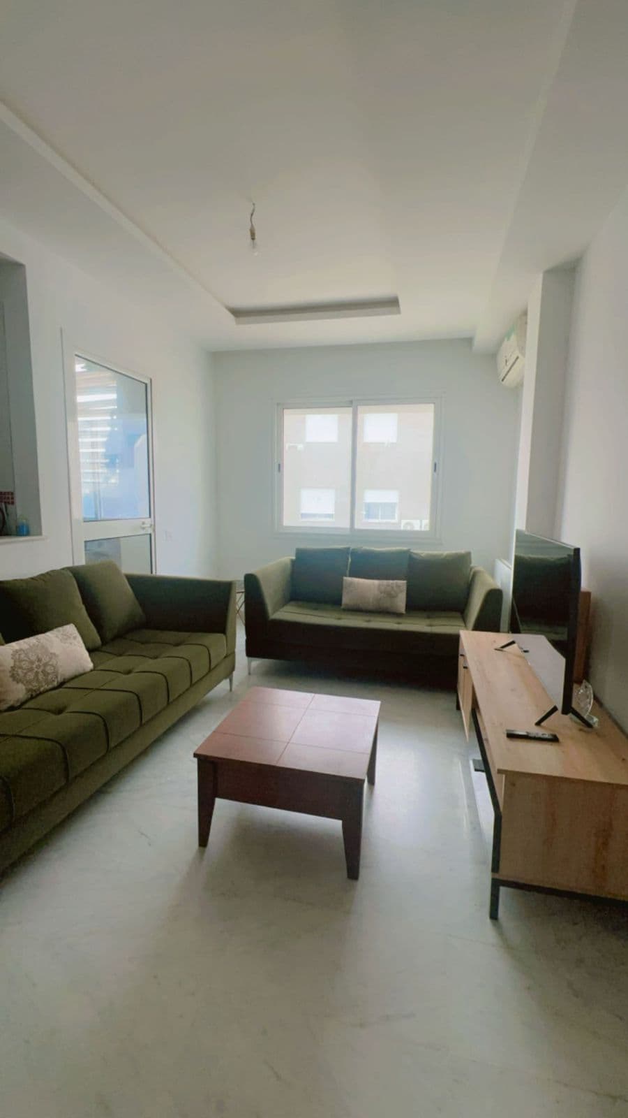 A bright living area features two green sofas arranged around a wooden coffee table. Natural light fills the room through two large windows, offering a view of the exterior. A television sits atop a light wooden stand against the wall.