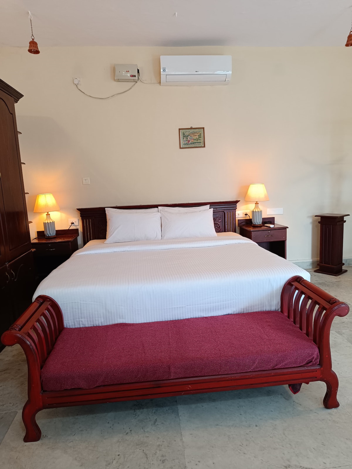 A cozy bedroom features a spacious bed with crisp white linens. Nightstands with lamps provide gentle lighting. A wooden bench with a maroon cushion sits at the foot of the bed. A wooden wardrobe and an air conditioning unit are visible on the walls.