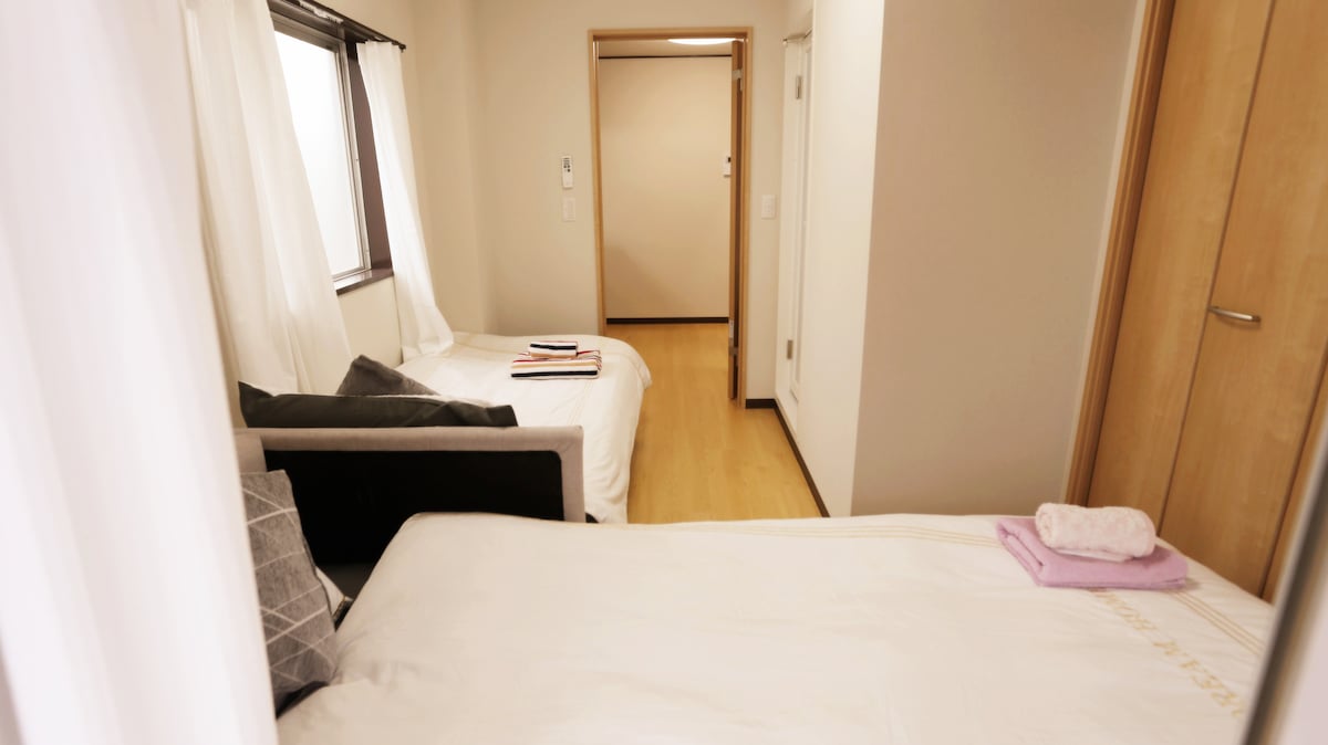 The room features two single beds arranged with bedding. Natural light enters through a window dressed in sheer curtains, illuminating the space. A small wall separates this area from the entrance hallway, which includes a closet and additional doorways.