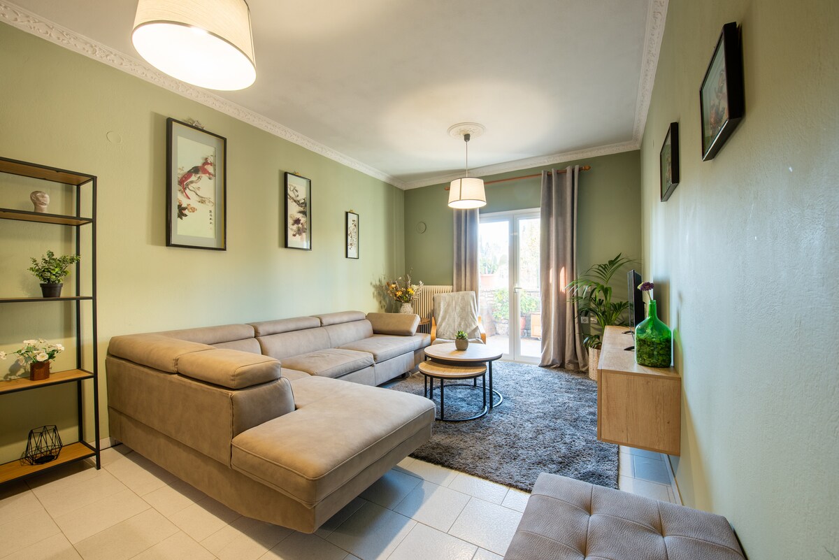 A living room features a spacious sectional sofa and a round coffee table, complemented by a plush area rug. Decorated with framed prints, the room is illuminated by natural light streaming in through sliding glass doors that offer access to a terrace.