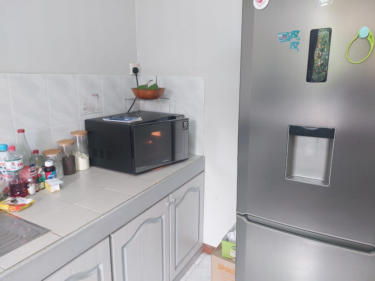 The kitchen features a modern fridge with an ice dispenser and a compact microwave atop the counter. A sink is visible alongside various containers and bottles, providing essential amenities for meal preparation and storage. Light tiles offer a clean backdrop.