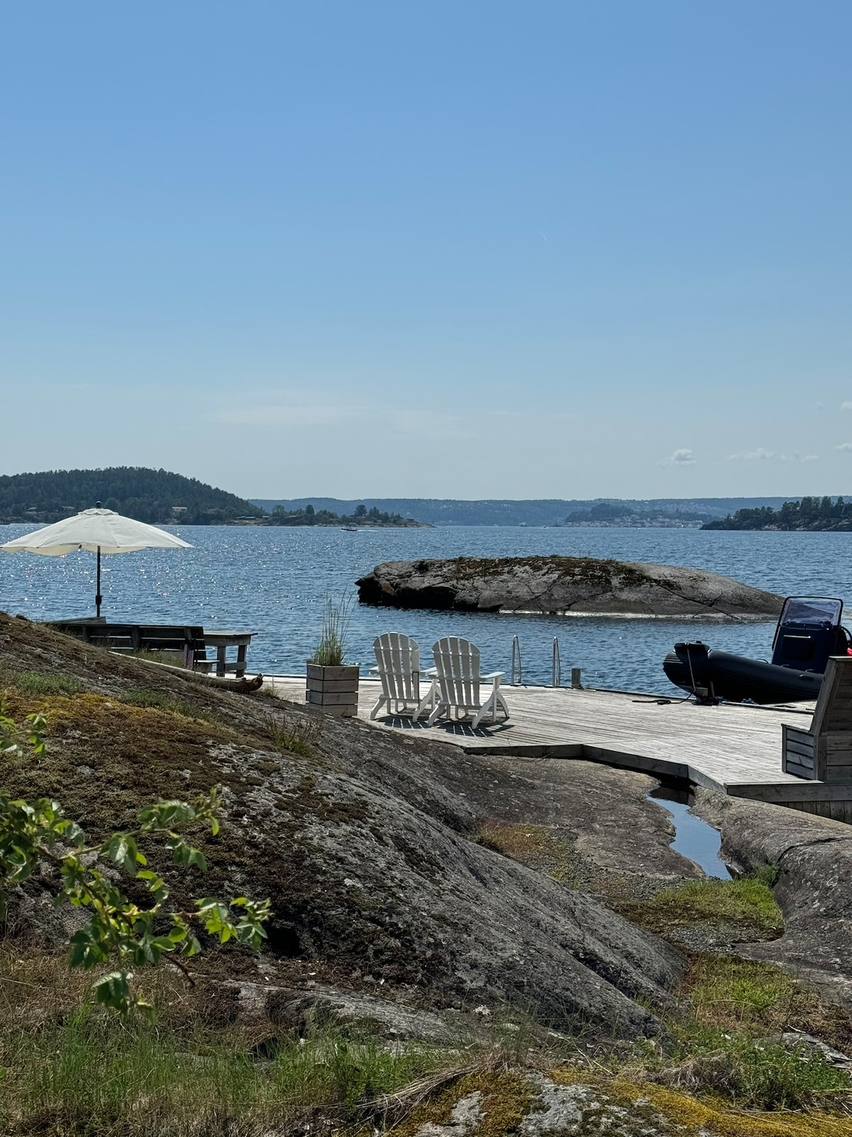 A wooden dock extends over calm waters, surrounded by rocky outcrops. Two white lounge chairs are positioned beside a large umbrella, offering shade. A small boat is visible nearby, with lush green hills in the distance under a clear blue sky.