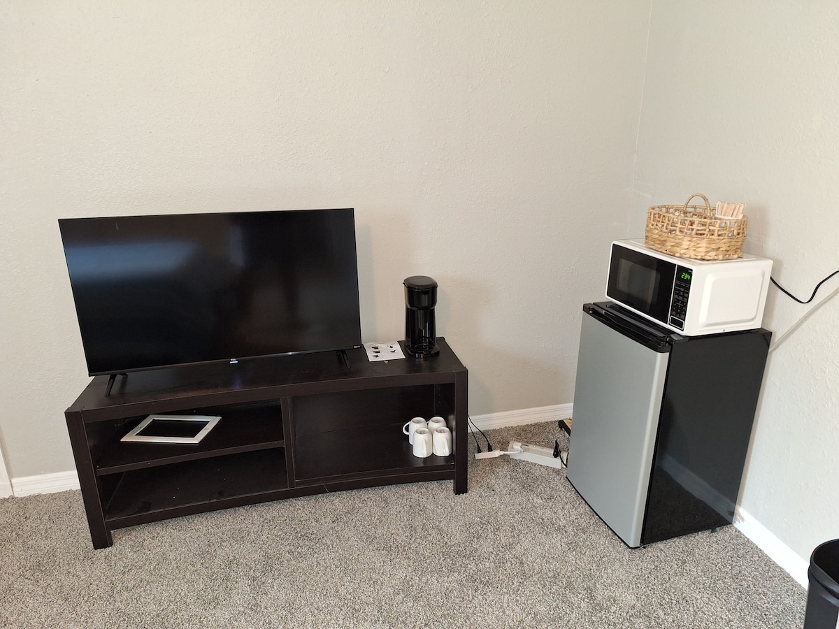 A compact living space features a flat-screen TV on a dark wooden stand, along with a coffee maker and a microwave placed beside a mini-fridge. A woven basket adds a touch of decor, while two cups are neatly arranged on the shelf below the TV.
