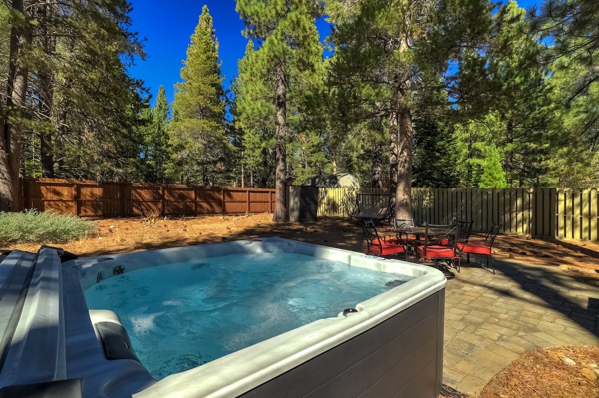 A bubbling hot tub is situated on a paver stone patio surrounded by tall pine trees, providing a sense of privacy. A small outdoor seating area with red cushioned chairs is visible, inviting relaxation in the serene backyard.