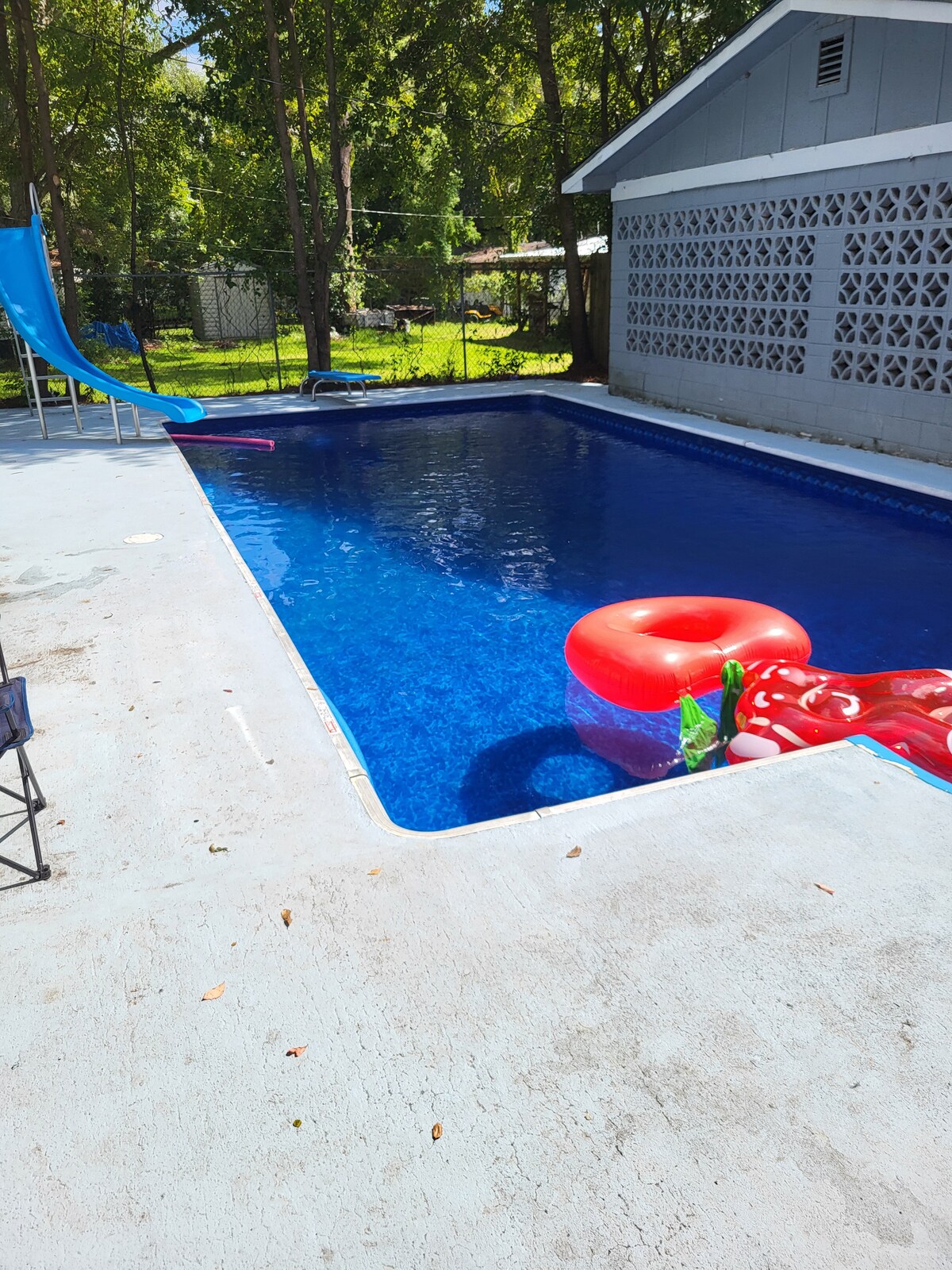 A well-maintained swimming pool is surrounded by a concrete patio. A bright blue slide leads into the water, which is inviting and clear. A large red float is positioned in the pool, adding a pop of color. Lush greenery provides a serene backdrop.