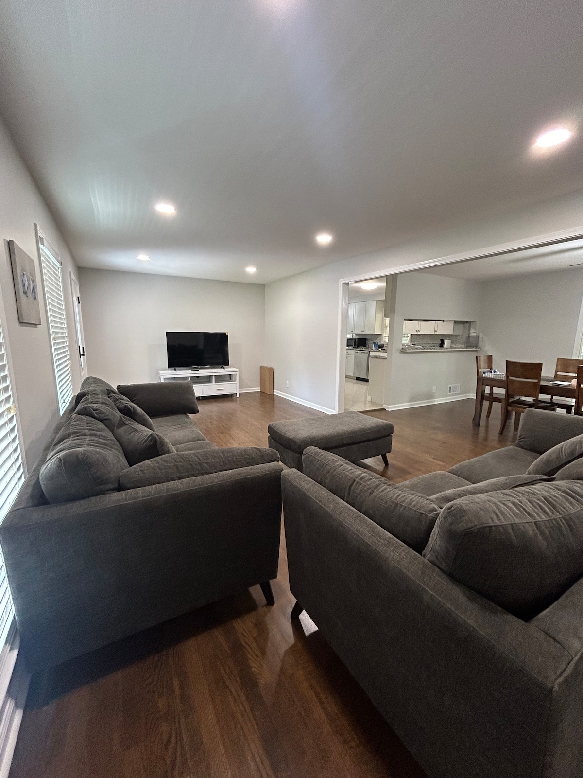 A spacious family room features a large, comfortable gray sectional and two armchairs. A coffee table sits centered within the seating area. A wall-mounted television is positioned on a white console, with soft lighting provided by recessed lights. The open layout connects to a dining area visible in the background.