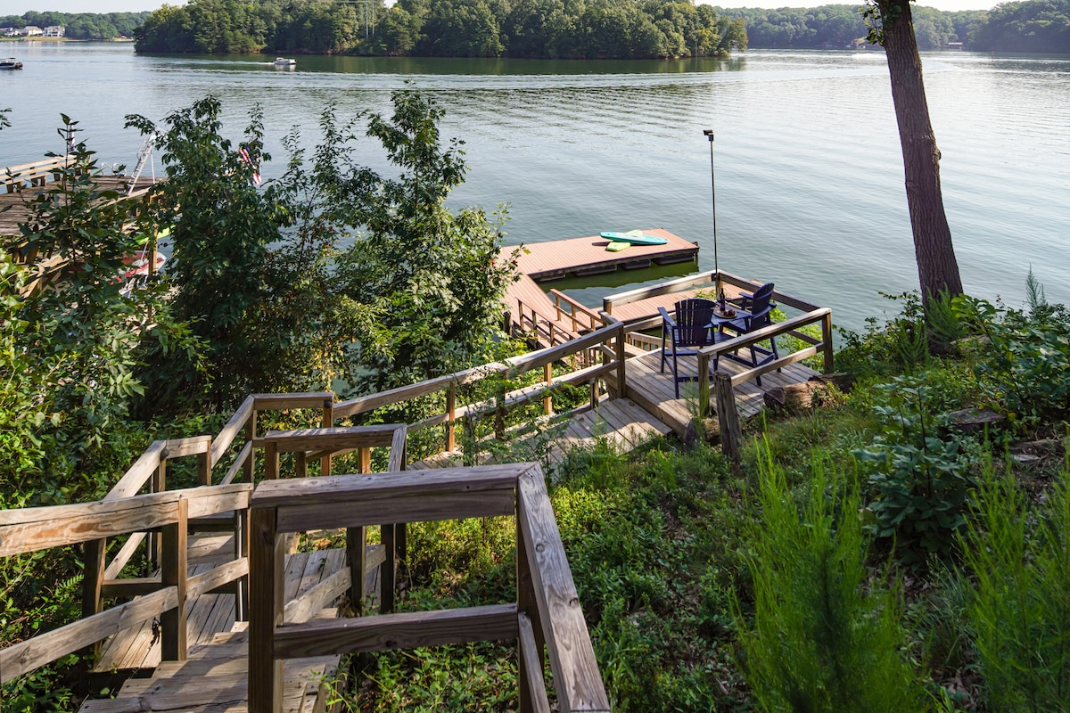 A wooden staircase winds down the hillside, leading to a private dock over tranquil waters. Adirondack chairs are positioned on the dock, surrounded by lush greenery and offering views of the calm lake and nearby shoreline.
