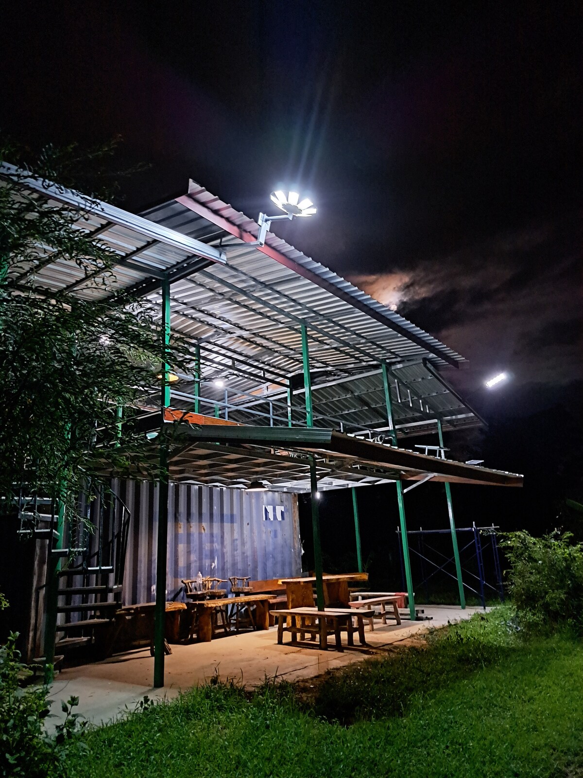 A two-story structure is shown, illuminated by bright lights at night. The building features a metal roof and elevated platform with green scaffolding. Below, a wooden dining table is visible, surrounded by chairs. Lush grass and foliage frame the scene.
