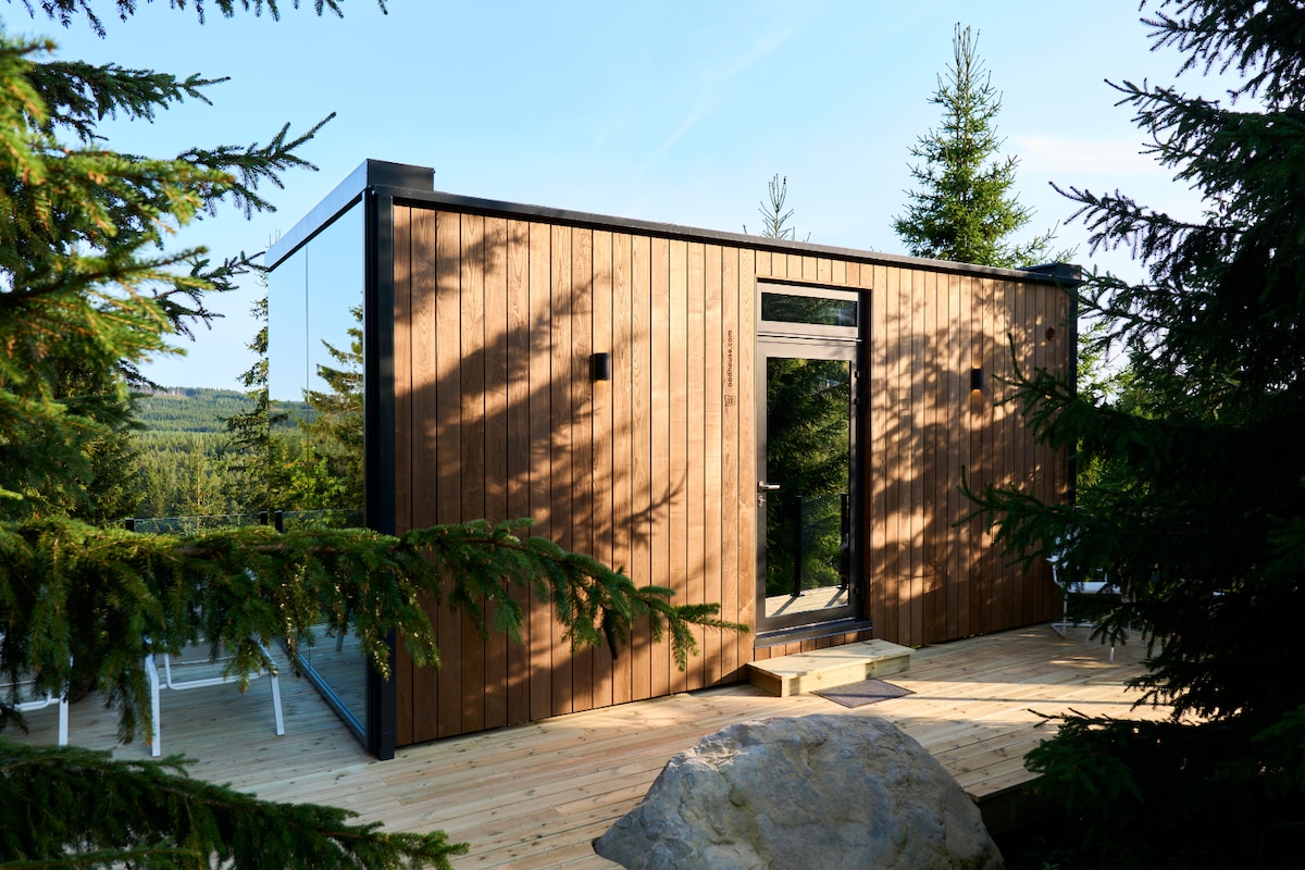 The cabin's exterior features a wooden facade with large glass panels, set amidst lush green trees. A wooden deck extends outward, providing a welcoming entryway illuminated by natural light. The surrounding trees create a serene privacy, enhancing the cabin's integration with nature.