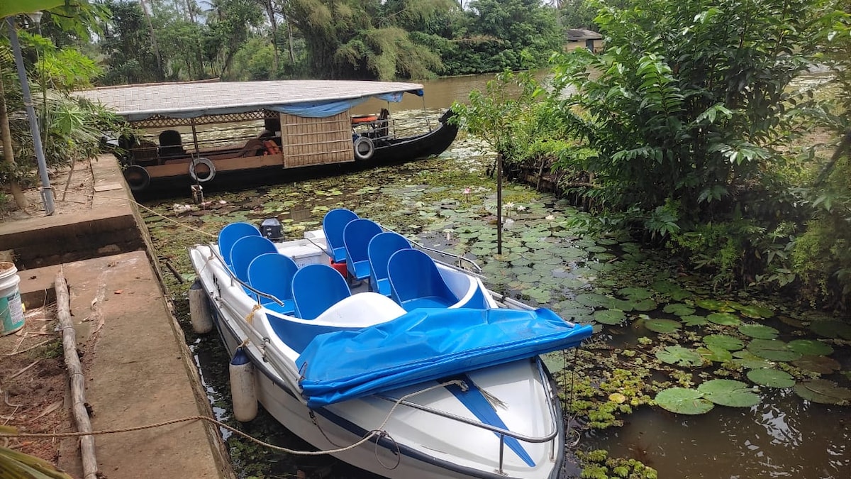 A motorboat with blue seating is anchored beside lush greenery and lily pads on the water's surface. In the background, a traditional boat is moored, with a wooden structure visible nearby. Natural vegetation frames the scene, enhancing the calmness of the setting.