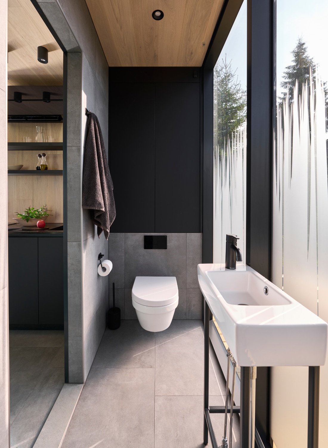 A modern bathroom is presented with sleek lines and a minimalist design. A compact toilet is positioned next to a stylish sink. Floor-to-ceiling glass enhances natural light, while a wood panel ceiling adds warmth. Shelving displays decor, contributing to the room's organized aesthetic.