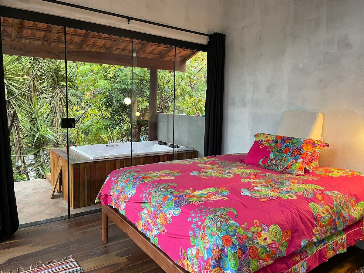 A spacious bedroom features a vibrant, floral-patterned bedding set on the bed. Large glass doors open to a serene outdoor area with a hot tub, surrounded by lush greenery. Natural light fills the room, enhancing the inviting atmosphere.