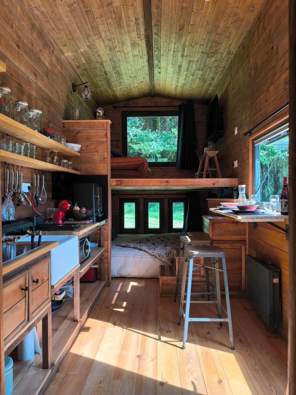 The interior of the tiny house features wooden walls and a high ceiling, creating a cozy vibe. A well-equipped kitchen area includes shelves filled with glassware. A sleeping loft can be seen above, accessible by a ladder. Large windows allow natural light to enter the space.