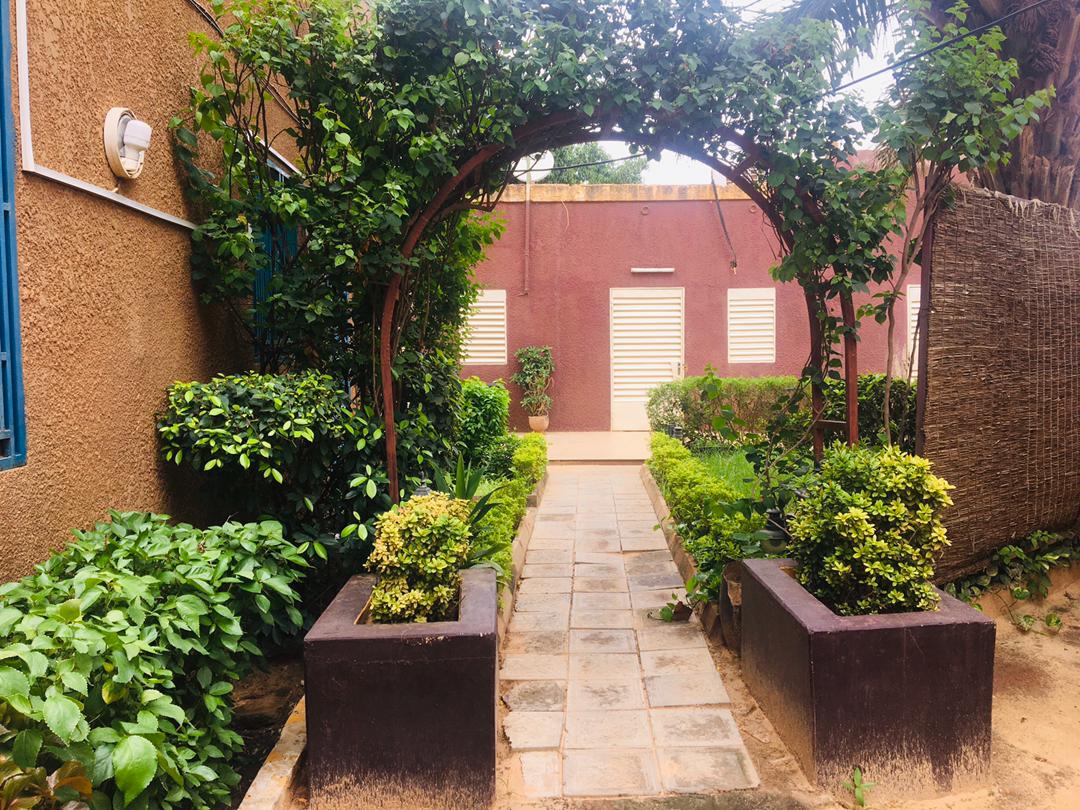 A landscaped pathway leads through a garden, framed by a curved wooden arch covered in greenery. Well-maintained bushes and flowering plants are displayed in planters on either side, guiding the way towards a building with closed shutters in the background.