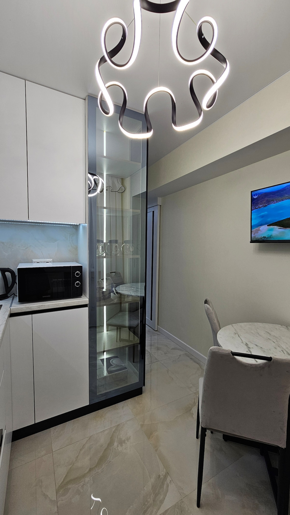 The modern kitchen area is highlighted by sleek cabinetry and a marble dining table with upholstered chairs. A stylish pendant light hangs above, while a wall-mounted TV and a glass partition reveal the nearby restroom. A microwave sits on the countertop.