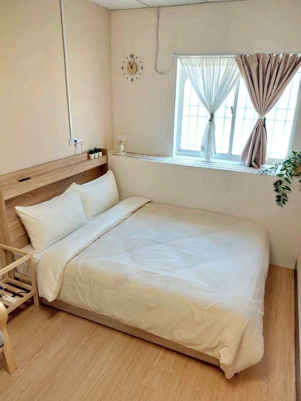 A bright sleeping area features a spacious bed with a simple white duvet and pillows. Natural light enters through a window adorned with light curtains, while a small clock and potted plant add a touch of decor. Light wood flooring complements the neutral tones.