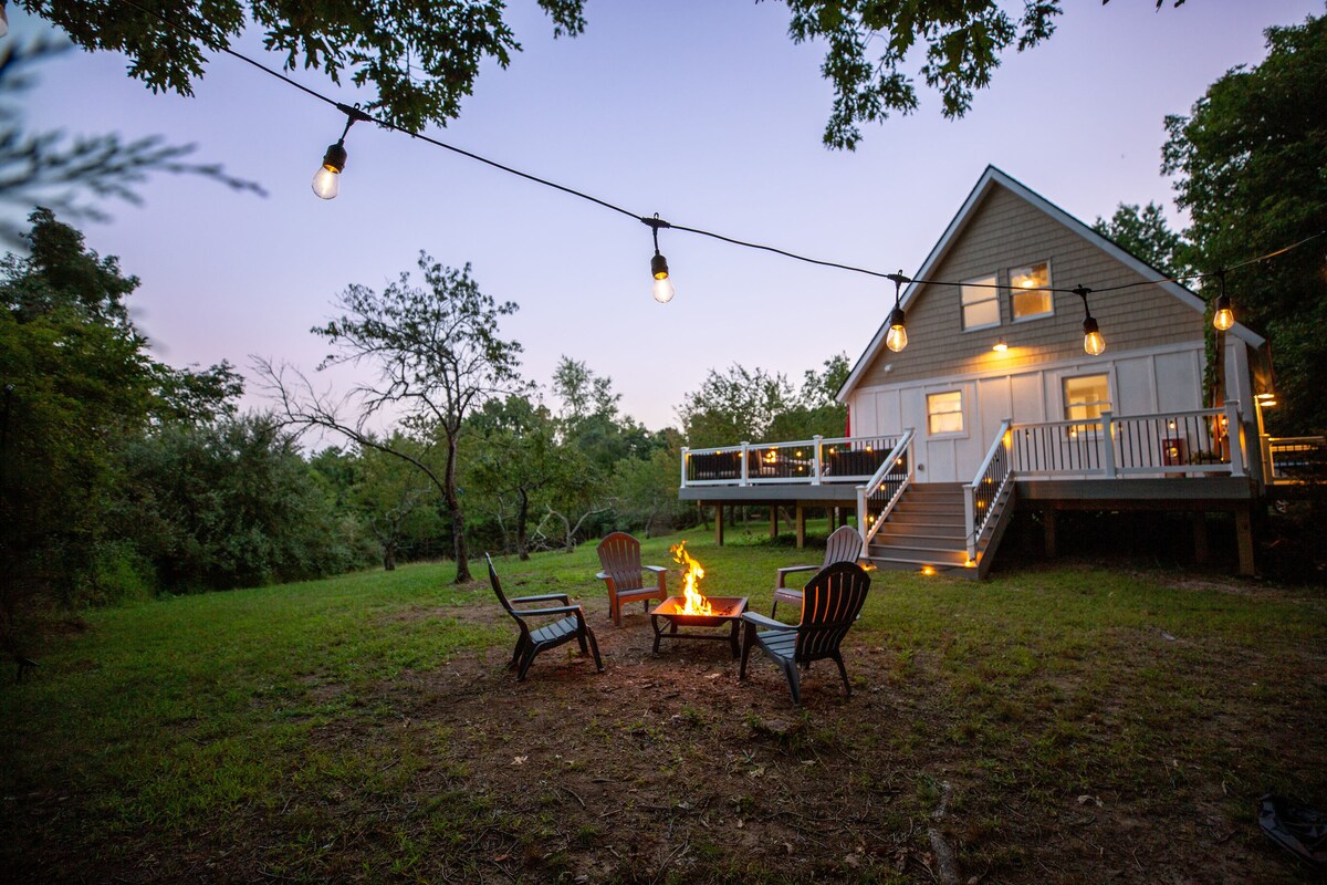 A charming cottage is illuminated by string lights, creating a welcoming atmosphere. A fire pit surrounded by several chairs is visible, set against a backdrop of lush greenery. The large deck extends from the cottage, providing a spacious area to enjoy the outdoors.