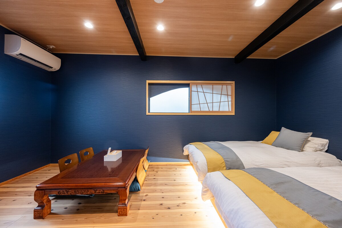 A contemporary room features two spacious beds with light bedding and soft throw pillows. A low wooden table with seating is positioned in the center, complemented by smooth wooden flooring. Soft lighting illuminates the space, while a large window provides natural light.