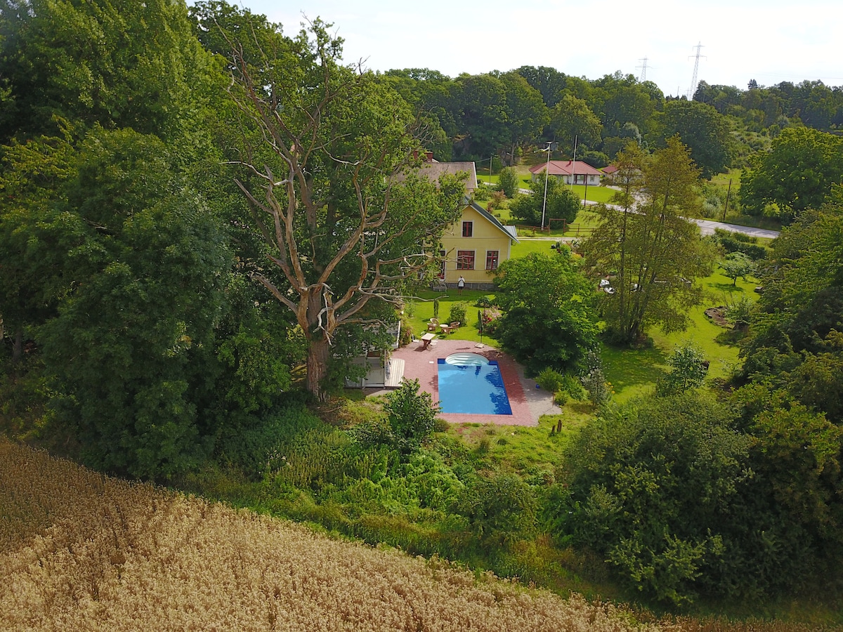 An aerial view showcases a tranquil garden oasis, featuring a swimming pool surrounded by greenery and seating areas. The large house, partially obscured by trees, offers a warm and inviting presence in the landscape, complemented by open grassy spaces and the surrounding fields.