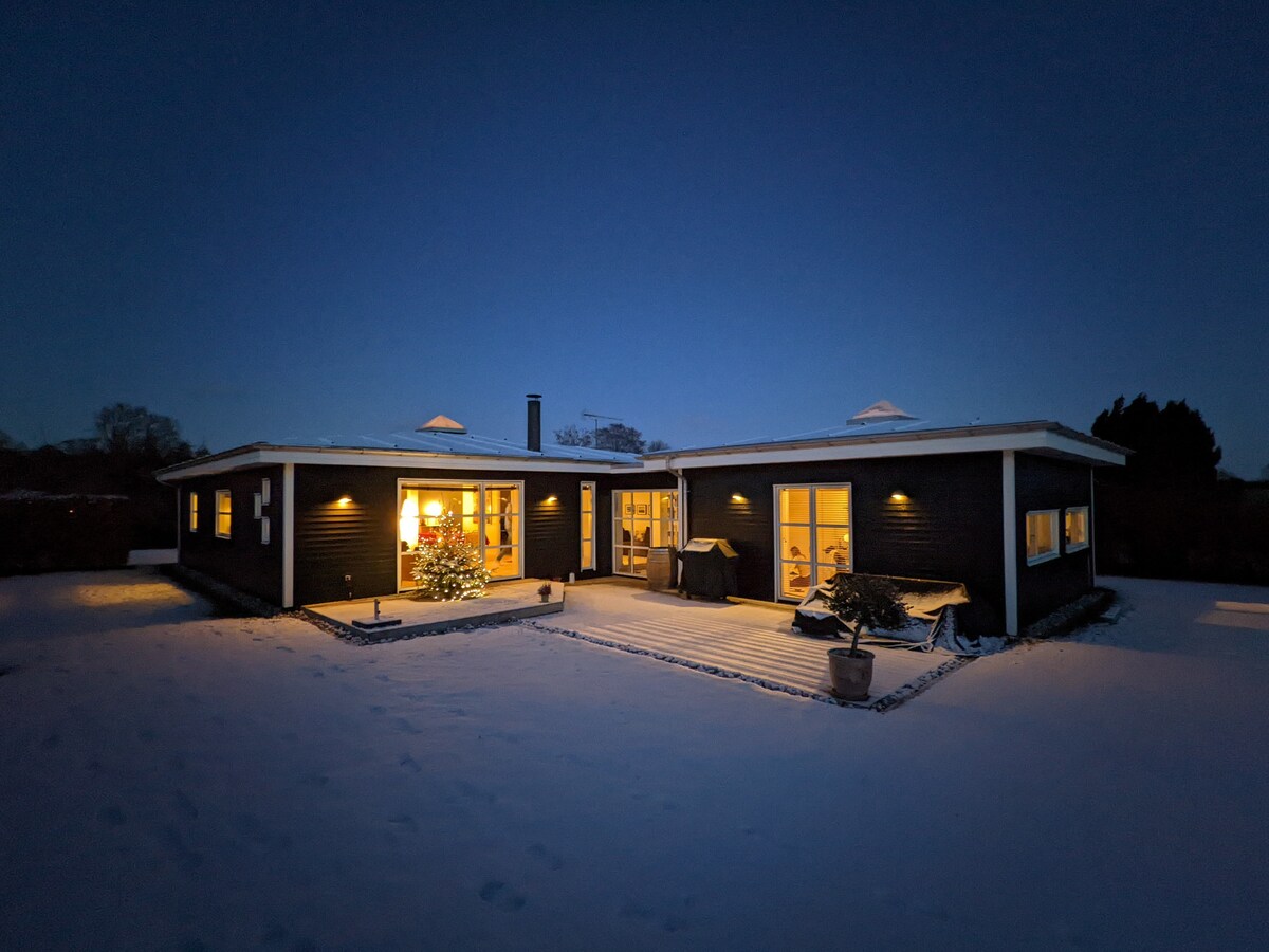 The modern home is illuminated against a twilight sky, surrounded by a snowy landscape. Large windows reveal a warm glow from within, highlighting the inviting entrance and a small evergreen tree on the covered porch. The architecture features a flat roof with ventilation skylights.