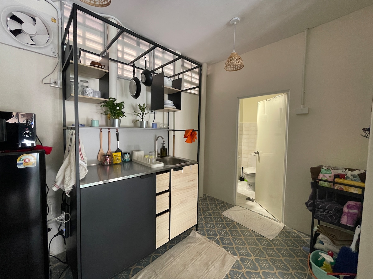 The kitchenette features a modern design with a metal and wood cabinetry. Cooking essentials are arranged on the shelves, complemented by plants for a touch of greenery. The space is well-lit, and a doorway leads to the bathroom area, visible through an open door.
