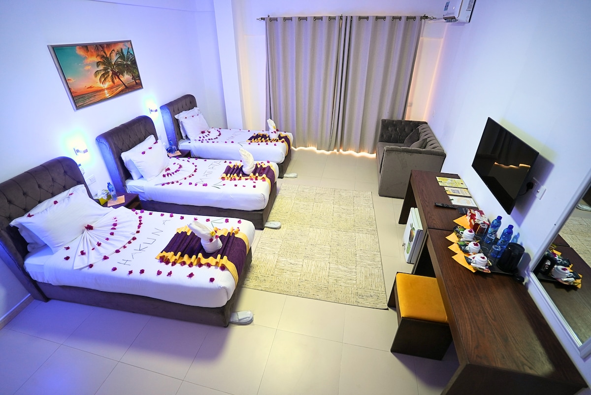 A spacious guest room is presented with three beds adorned with decorative linens and rose petals. A plush seating area is provided next to the window, while a modern desk with assorted beverages and amenities is featured. Soft lighting enhances the welcoming ambiance.