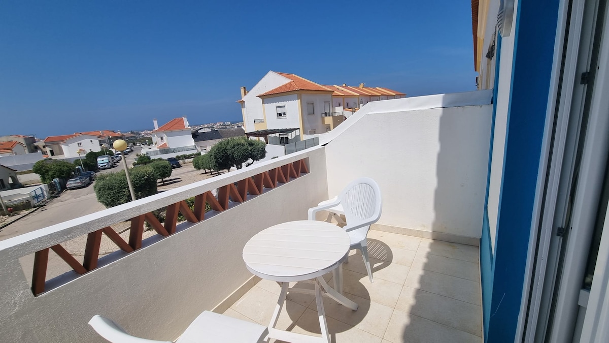 A private terrace is presented with a small round table and two white chairs. The surrounding area includes a view of nearby buildings and a clear blue sky. The space is bordered by a low white wall, enhancing the openness of the outdoor setting.