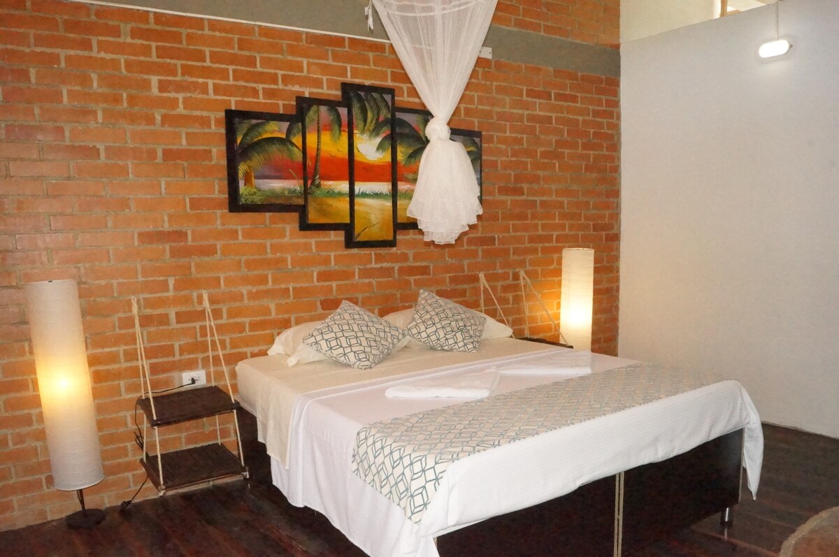 A spacious bedroom features a large bed with decorative pillows, positioned against a textured brick wall. Artistic wall hangings depicting tropical themes are displayed above the bed, while illuminated floor lamps provide soft lighting on either side.