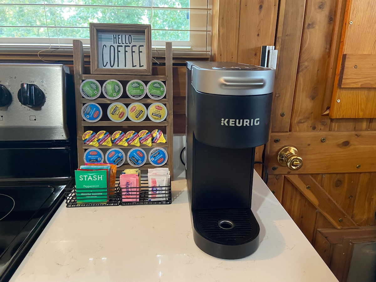 A coffee station features a black Keurig machine alongside a display rack filled with an assortment of coffee pods. A small sign reading 'Hello Coffee' is presented above the setup. The countertop reflects a clean and organized space, with wooden cabinetry in the background.