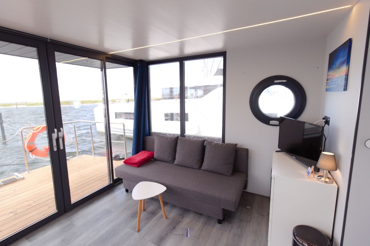 A modern living area features large glass doors that offer views of the water. A gray sofa is complemented by a small, round table and a television. A decorative mirror and a lamp enhance the space, with natural light flowing in through the windows.