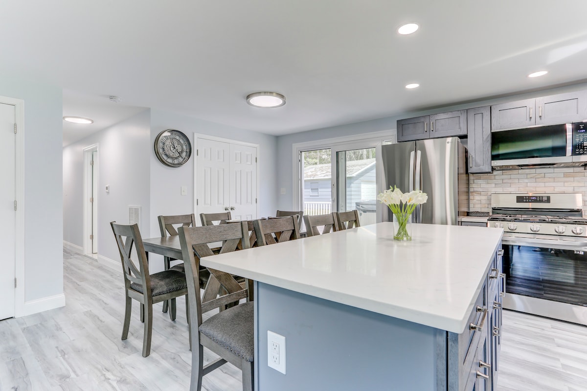 A modern kitchen and dining area is featured, with a large central island and six dining chairs surrounding a rectangular table. Stainless steel appliances, including a refrigerator and stove, are visible, complemented by pale cabinetry and bright lighting.