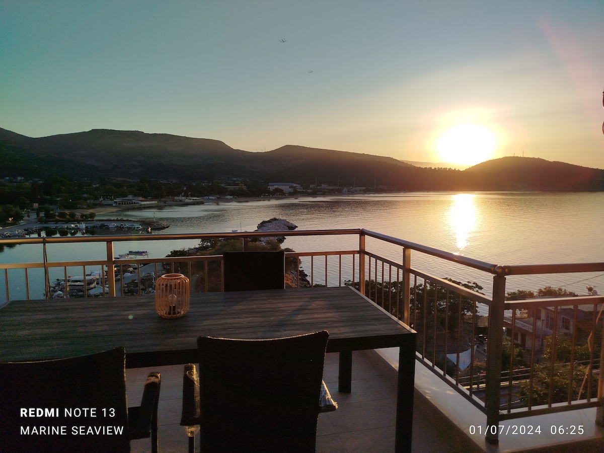 A balcony view showcases a serene sunrise over the sea, with mountains in the background. A table and two chairs are positioned on the balcony, providing a perfect spot for relaxation and enjoying the landscape.